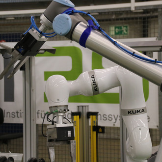 Foto eines KUKA-Roboterarms in einem industriellen Umfeld. Der Roboterarm ist weiß mit blauen Gelenken und trägt die Aufschrift "KUKA" an mehreren Stellen. Er hält ein Werkzeug oder Gerät am Ende seines Arms. Im Hintergrund ist ein Schild mit dem teilweise verdeckten Logo des Instituts für Produktionssysteme (IPS) zu sehen, wobei das "I" grau und das "P" und "S" grün gefärbt sind. Der Roboter befindet sich in einer Werkstattumgebung mit verschiedenen technischen Geräten und Sicherheitsbarrieren im Hintergrund.