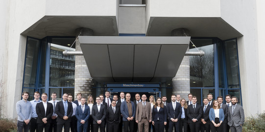 Foto einer großen Gruppe von Menschen in Geschäftskleidung, die vor einem Gebäude mit der Aufschrift "Maschinenbau" stehen. Die Gruppe besteht aus Männern und Frauen, die in zwei Reihen nebeneinander posieren. Das Gebäude im Hintergrund hat eine moderne Architektur mit großen Fenstern und einem breiten Vordach. Der Boden vor dem Gebäude ist mit Kopfsteinpflaster bedeckt. Die Szene wirkt wie ein formelles Gruppenfoto, möglicherweise von einer Abschlussklasse oder einem Team in einem beruflichen Kontext.
