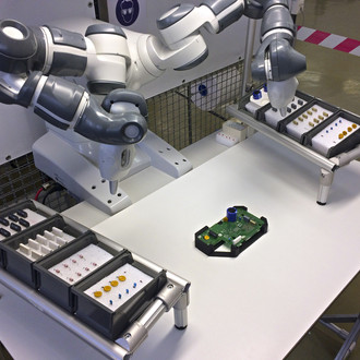 Foto: Ein Roboterarm arbeitet an einem weißen Tisch. Der Tisch ist mit verschiedenen Tabletts bestückt, die kleine elektronische Bauteile enthalten. In der Mitte des Tisches befindet sich eine grüne Leiterplatte mit verschiedenen Komponenten. Der Roboterarm scheint präzise an der Leiterplatte zu arbeiten. Im Hintergrund sind Teile einer industriellen Umgebung zu erkennen, darunter ein mit rot-weißem Absperrband markierter Bereich.