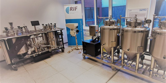 Foto eines Labors mit industriellen Anlagen. Links steht eine komplexe Apparatur aus Metall mit verschiedenen Ventilen und Rohrleitungen, neben der ein Bildschirm angebracht ist. Rechts befinden sich drei große, glänzende Edelstahlbehälter auf Rollen, die durch Rohrleitungen miteinander verbunden sind. Im Hintergrund ist ein Banner mit der Aufschrift "RIF" und ein Fenster mit Blick nach draußen zu sehen.