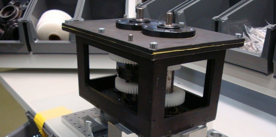 Foto eines mechanischen Aufbaus auf einer Arbeitsfläche. Im Vordergrund befindet sich eine Vorrichtung, die aus einem stabilen, rechteckigen Rahmen besteht, der Zahnräder und mechanische Komponenten enthält. Die Vorrichtung ist auf einer perforierten Metallplatte montiert, die zur Befestigung auf der Arbeitsfläche dient. Rechts und links der Vorrichtung sind Behälter mit verschiedenen Schrauben und mechanischen Teilen zu sehen. Im Hintergrund befinden sich weitere Ablagefächer mit großen weißen Zahnrädern und anderen Bauteilen. Die gesamte Anordnung deutet auf einen Arbeitsplatz hin, der für die Montage oder Wartung mechanischer Systeme eingerichtet ist.