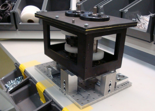 Foto eines mechanischen Aufbaus auf einer Arbeitsfläche. Im Vordergrund befindet sich eine Vorrichtung, die aus einem stabilen, rechteckigen Rahmen besteht, der Zahnräder und mechanische Komponenten enthält. Die Vorrichtung ist auf einer perforierten Metallplatte montiert, die zur Befestigung auf der Arbeitsfläche dient. Rechts und links der Vorrichtung sind Behälter mit verschiedenen Schrauben und mechanischen Teilen zu sehen. Im Hintergrund befinden sich weitere Ablagefächer mit großen weißen Zahnrädern und anderen Bauteilen. Die gesamte Anordnung deutet auf einen Arbeitsplatz hin, der für die Montage oder Wartung mechanischer Systeme eingerichtet ist.