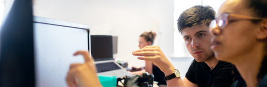 Foto zweier Personen, die intensiv an einem Computerbildschirm arbeiten. Der Mann links zeigt auf den Bildschirm und diskutiert etwas mit der Frau rechts, die eine Brille trägt und ebenfalls auf den Bildschirm schaut. Im Hintergrund arbeitet eine weitere Person an einem Laptop. Der Raum ist hell und modern eingerichtet. Die Szene vermittelt den Eindruck einer konzentrierten Zusammenarbeit in einer Büroumgebung.