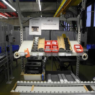 Foto eines Arbeitsplatzes in einer Fabrik, der für die Gehäusemontage eingerichtet ist. Auf dem Tisch sind verschiedene Behälter mit Kleinteilen und Komponenten zu sehen, darunter schwarze Gehäuseteile und rote Kästchen mit Schrauben. Oberhalb des Arbeitsplatzes hängt ein Schild mit der Aufschrift "AP 1 Gehäusemontage". Im Hintergrund sind weitere industrielle Arbeitsbereiche und Ausrüstungen zu erkennen.
