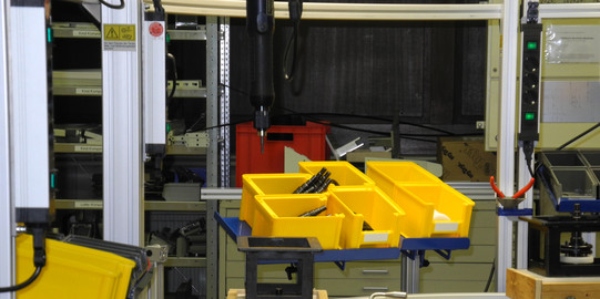 Foto einer automatisierten Fertigungsanlage. Im Vordergrund sind mehrere gelbe Behälter auf einem Förderband zu sehen, die verschiedene Bauteile enthalten. Die Anlage ist von metallenen Stützen und Leitungen umgeben, im Hintergrund sind weitere Maschinen und Werkzeuge zu erkennen.