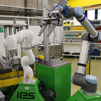 Das Bild zeigt eine industrielle Arbeitsstation mit zwei Roboterarmen. Der linke Roboterarm ist weiß und der rechte Roboterarm ist blau und silber. Beide Roboter sind auf grünen Maschinen mit der Aufschrift "IPS" montiert. Im Hintergrund sind Regale mit verschiedenen Werkzeugen und Materialien sowie weitere industrielle Ausrüstungen zu sehen. Die Arbeitsstation befindet sich in einem gut organisierten und sauber gehaltenen industriellen Umfeld.