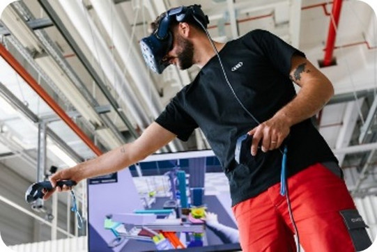 Foto, das eine Person in einer industriellen Umgebung zeigt, die ein Virtual-Reality-Headset trägt und mit zwei VR-Controllern interagiert. Die Person steht in einem technisch ausgestatteten Raum mit sichtbaren Leitungen und Beleuchtungselementen an der Decke. Hinter ihr befindet sich ein Bildschirm, auf dem ein digitales Modell einer Produktionsanlage dargestellt ist, das offensichtlich mit der VR-Anwendung verknüpft ist. Die Körperhaltung und Armbewegung der Person deuten darauf hin, dass sie innerhalb der virtuellen Umgebung einen Arbeitsprozess oder ein Anlagenlayout simuliert. Das Bild vermittelt den Einsatz von VR-Technik zur Analyse, Planung oder Schulung im industriellen Kontext.