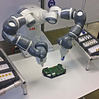 Foto eines ABB-Roboterarms in einer Produktionsumgebung. Der duale Roboterarm hat graue und weiße Gelenke und bearbeitet eine elektronische Platine, die auf einem Tisch vor ihm liegt. Links und rechts vom Roboter befinden sich Ablagen mit sortierten Bauteilen. Der Hintergrund besteht aus einem Gitter und weiteren industriellen Elementen.