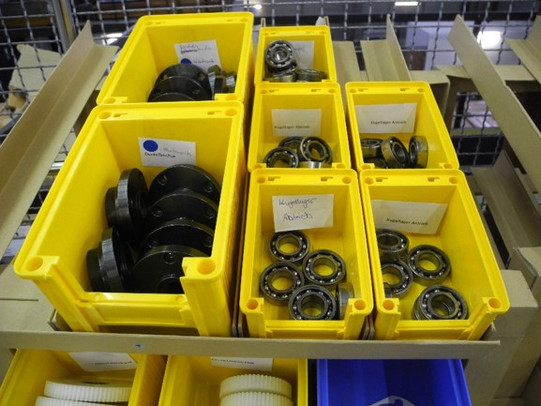 Das Bild zeigt eine Reihe von gelben Kunststoffbehältern, die verschiedene Kugellager und Metallteile enthalten. Die Behälter sind ordentlich auf einem Regal oder einer Werkbank angeordnet. In jedem Behälter sind die Teile sauber gestapelt und durch Beschriftungen gekennzeichnet, wie zum Beispiel "Kugellager Innen" und "Kugellager Außen". Diese Beschriftungen helfen bei der schnellen Identifikation der Teile. Im Hintergrund sind weitere Regale und möglicherweise andere Arbeitsbereiche zu sehen. Die Umgebung wirkt gut organisiert und strukturiert, was auf eine effiziente Arbeitsweise hinweist.