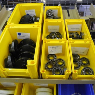 Das Bild zeigt eine Reihe von gelben Kunststoffbehältern, die verschiedene Kugellager und Metallteile enthalten. Die Behälter sind ordentlich auf einem Regal oder einer Werkbank angeordnet. In jedem Behälter sind die Teile sauber gestapelt und durch Beschriftungen gekennzeichnet, wie zum Beispiel "Kugellager Innen" und "Kugellager Außen". Diese Beschriftungen helfen bei der schnellen Identifikation der Teile. Im Hintergrund sind weitere Regale und möglicherweise andere Arbeitsbereiche zu sehen. Die Umgebung wirkt gut organisiert und strukturiert, was auf eine effiziente Arbeitsweise hinweist.