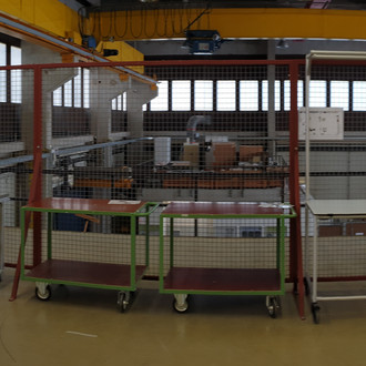 Panoramafoto: Eine weitläufige industrielle Werkstatt mit mehreren Arbeitsbereichen und Regalen. Links befinden sich Schreibtische und Regale mit Büromaterialien. In der Mitte sind mehrere Arbeitsstationen mit Montagetischen und Werkzeugen angeordnet. Rechts sind weitere Maschinen und Arbeitsgeräte zu sehen. Die Werkstatt ist hell beleuchtet durch große Fenster im oberen Bereich und verfügt über eine gelbe Laufkatze an der Decke für den Transport von schweren Lasten. Der Raum wirkt gut organisiert und geräumig.