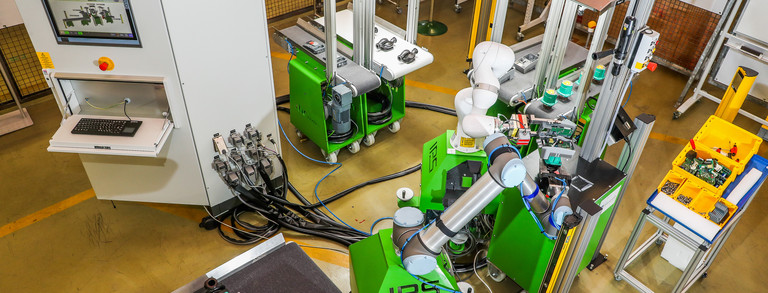 Foto einer automatisierten Produktionslinie in einer Fabrikhalle, aufgenommen von oben. Im Zentrum des Bildes befindet sich ein grüner Roboterarm, der von mehreren grünen Maschinen und Förderbändern umgeben ist. Rechts sind gelbe Behälter mit verschiedenen Kleinteilen zu sehen. Links steht ein weißer Kontrollschrank mit einem Bildschirm und einer Tastatur. Die Szene zeigt eine hochmoderne Fertigungsumgebung mit mehreren miteinander verbundenen Maschinen, die durch den Roboterarm gesteuert werden. Kabel und Leitungen verlaufen über den Boden, was auf die Komplexität der Anlage hinweist.