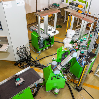Foto einer automatisierten Produktionslinie in einer Fabrikhalle, aufgenommen von oben. Im Zentrum des Bildes befindet sich ein grüner Roboterarm, der von mehreren grünen Maschinen und Förderbändern umgeben ist. Rechts sind gelbe Behälter mit verschiedenen Kleinteilen zu sehen. Links steht ein weißer Kontrollschrank mit einem Bildschirm und einer Tastatur. Die Szene zeigt eine hochmoderne Fertigungsumgebung mit mehreren miteinander verbundenen Maschinen, die durch den Roboterarm gesteuert werden. Kabel und Leitungen verlaufen über den Boden, was auf die Komplexität der Anlage hinweist.