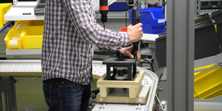 Ein Mann in einem karierten Hemd steht an einem Förderband und verwendet ein Elektrowerkzeug, um eine Baugruppe zusammenzusetzen. Im Hintergrund befinden sich gelbe und blaue Behälter mit verschiedenen Bauteilen.