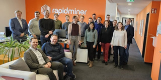 Foto: Eine Gruppe von 18 Personen steht und sitzt in einem Büro vor einer orangefarbenen Wand mit dem weißen Logo und Schriftzug "rapidminer". Die Personen lächeln in die Kamera und tragen überwiegend legere Kleidung. Im Vordergrund sitzen zwei Personen auf einem weißen Sofa, während die restlichen Personen im Hintergrund stehen. Der Raum ist modern eingerichtet und hell beleuchtet.