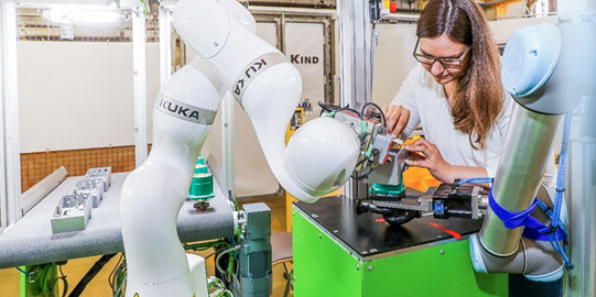 Foto einer Frau, die in einer industriellen Umgebung an einer Maschine arbeitet. Die Frau trägt eine Brille und konzentriert sich auf ein Bauteil, das sie bearbeitet. Neben ihr befinden sich zwei Roboterarme der Marke KUKA, die an einer grünen Arbeitsstation befestigt sind. Im Hintergrund sind weitere industrielle Ausrüstungen und eine helle, gut beleuchtete Werkstattumgebung zu sehen.