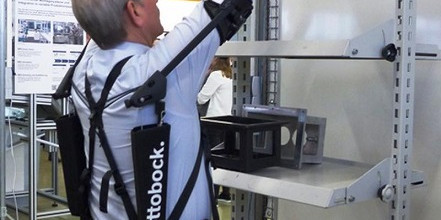 Das Bild zeigt einen Mann mit grauen Haaren und Brille, der ein Exoskelett der Marke "Ottobock" trägt. Er steht vor einem Metallregal und hebt ein Objekt in das Regal. Das Exoskelett ist schwarz und unterstützt die Arme und den Rücken des Mannes. Im Hintergrund sind weitere industrielle Arbeitsplätze und Geräte zu sehen.