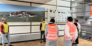 Besuchergruppe bei einer Werksführung mit Schautafel und Produktionsplan.