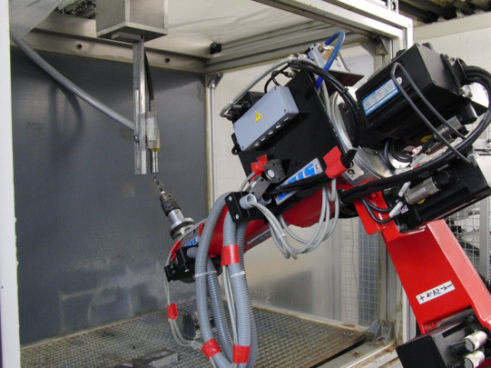 Foto eines Industrieroboters in einer Arbeitszelle. Der Roboterarm ist rot und hat mehrere Gelenke sowie Kabel und Schläuche, die ihn mit Strom und Daten versorgen. Der Roboter hält ein Werkzeug, das auf ein Metallteil in einer geschlossenen Kammer ausgerichtet ist. Die Arbeitszelle ist aus Metall und Glas gefertigt und dient dem Schutz sowie der Kontrolle des Arbeitsprozesses. Die gesamte Szene deutet auf einen automatisierten Fertigungsprozess hin, bei dem Präzision und Sicherheit im Vordergrund stehen.