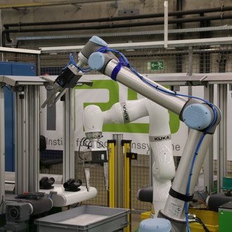 Foto eines KUKA-Roboterarms in einer industriellen Umgebung. Der Roboterarm ist weiß mit blauen Akzenten und mehreren Gelenken, die eine flexible Bewegung ermöglichen. Der Greifer am Ende des Arms hält ein Werkzeug oder Bauteil. Im Hintergrund sind industrielle Geräte, Sicherheitszäune und ein großes Banner mit der Aufschrift "Institut für Produktionssysteme" zu sehen. Die Szene befindet sich in einer modernen Fertigungs- oder Forschungsanlage, die auf fortschrittliche Automatisierung und Robotik spezialisiert ist. Der Roboterarm scheint Teil eines automatisierten Prozesses oder einer technischen Demonstration zu sein.