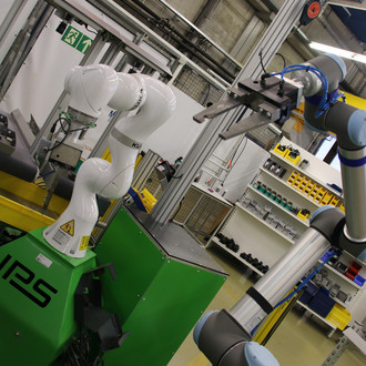 Das Bild zeigt eine industrielle Umgebung mit zwei Roboterarmen, die an grünen Maschinen montiert sind. Der linke Roboterarm ist weiß und der rechte Roboterarm ist blau und silber. Beide Roboter sind in einer Arbeitsstation positioniert, umgeben von Regalen mit Werkzeugen und Materialien. Im Hintergrund sind metallene Strukturen und elektrische Kabel zu sehen. Auf der linken grünen Maschine ist der Schriftzug "IPS" sichtbar.