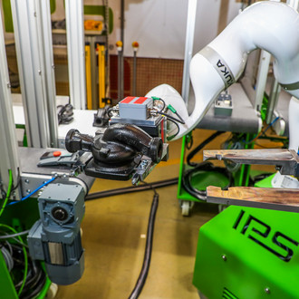 Das Bild zeigt eine industrielle Arbeitsstation mit einem weißen Roboterarm der Marke KUKA, der an einer grünen Maschine mit der Aufschrift "IPS" montiert ist. Der Roboterarm hält ein mechanisches Bauteil mit einer Greifvorrichtung. Um die Maschine herum sind verschiedene technische Geräte und Verkabelungen sichtbar. Im Hintergrund befinden sich weitere Maschinen und industrielle Ausrüstungen.