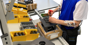 Foto eines Arbeiters, der an einem Montagearbeitsplatz arbeitet. Der Arbeiter trägt ein blau-weißes Adidas-Shirt und hat mehrere Sensoren an seinen Armen angebracht, die mit einem kleinen Gerät an seinem Gürtel verbunden sind. Er benutzt ein rotes Werkzeug, um an einem mechanischen Bauteil zu arbeiten. Auf dem Arbeitstisch sind mehrere gelbe und graue Behälter mit Schrauben und anderen Kleinteilen zu sehen. Im Hintergrund befinden sich weitere Arbeitsmaterialien und Geräte. Die Szene zeigt eine moderne Produktionsumgebung mit Fokus auf Ergonomie und Datenerfassung.