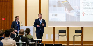 Foto einer Präsentation in einem Konferenzraum. Zwei Personen, ein Mann und eine Frau, stehen vorne und halten eine Präsentation. Beide tragen Geschäftskleidung. Der Mann spricht und gestikuliert, während die Frau neben einem Podium steht. Hinter ihnen ist eine große Leinwand, auf der eine Folie mit einem Bild eines Tablets und einem Waschmaschinenbild sowie Textinhalten zu sehen ist. Im Vordergrund sitzen mehrere Zuhörer an Tischen und verfolgen die Präsentation. Der Raum ist modern eingerichtet und gut beleuchtet.