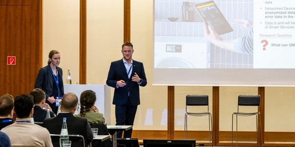 Foto einer Präsentation in einem Konferenzraum. Zwei Personen, ein Mann und eine Frau, stehen vorne und halten eine Präsentation. Beide tragen Geschäftskleidung. Der Mann spricht und gestikuliert, während die Frau neben einem Podium steht. Hinter ihnen ist eine große Leinwand, auf der eine Folie mit einem Bild eines Tablets und einem Waschmaschinenbild sowie Textinhalten zu sehen ist. Im Vordergrund sitzen mehrere Zuhörer an Tischen und verfolgen die Präsentation. Der Raum ist modern eingerichtet und gut beleuchtet.
