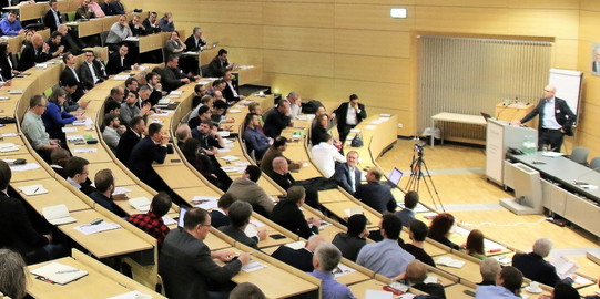Foto eines vollen Hörsaals während einer Vorlesung oder Konferenz. Viele Zuhörer sitzen in den ansteigenden Sitzreihen, einige machen sich Notizen oder verfolgen die Präsentation. Vorne rechts steht ein Redner an einem Pult und spricht zu den Anwesenden. Ein Kamerastativ ist ebenfalls zu sehen, was darauf hindeutet, dass die Veranstaltung aufgezeichnet wird. Der Raum hat helle Holzverkleidungen und wirkt gut beleuchtet.