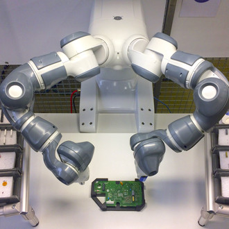 Foto eines dualen Roboterarms in einer Produktionsumgebung. Der Roboterarm hat zwei Gelenke, die jeweils mit grauen und weißen Segmenten ausgestattet sind. Vor dem Roboterarm befindet sich eine elektronische Platine, die auf einem Tisch liegt. Links und rechts davon sind mehrere Ablagen mit sortierten Bauteilen zu sehen. Der Hintergrund besteht aus einem Gitter und weiteren industriellen Elementen.