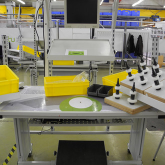 Foto eines industriellen Arbeitsplatzes. Auf dem Tisch befinden sich gelbe Behälter, ein schwarzes Fach, eine weiße Arbeitsunterlage und ein Halter mit mehreren Werkzeugen. Über dem Tisch ist ein Monitor angebracht, der von einer Metallkonstruktion gehalten wird. Im Hintergrund sind weitere Arbeitsstationen, Regale mit blauen Behältern und industrielle Ausrüstung zu sehen. Der Raum ist gut beleuchtet, und der Boden ist hell.