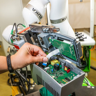 Nahaufnahme einer Hand, die an einer offenen elektronischen Baugruppe arbeitet. Die Baugruppe ist in einem Gehäuse montiert und enthält grüne Leiterplatten und verschiedene elektronische Komponenten. Im Hintergrund ist ein weißer KUKA-Roboterarm zu sehen, der möglicherweise bei der Montage hilft. Die Szene spielt in einer industriellen Umgebung mit weiteren Maschinen und Werkbänken im Hintergrund.