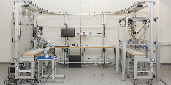 U-förmige Montagelinie aus Aluminiumprofilen in der IETC-Laborfläche. Links steht ein Roboterarm, rechts ein Deltaroboter. In der Mitte befinden sich ein Bildschirm, Werkstückträger auf einem Transportband und oberhalb davon eine Prüfstation.