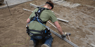 A worker kneels on a construction site wearing an exoskeleton. Tools are laid out on the sandy ground in front of him, and paved areas with construction materials are visible in the background.