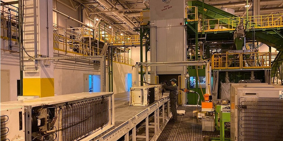 Foto einer Industriehalle mit moderner Recyclinganlage. Im Vordergrund sind mehrere Förderbänder und Maschinen zu sehen. Ein Arbeiter bedient eine der Maschinen, während im Hintergrund eine komplexe Struktur aus Geländern, Treppen und weiteren Förderbändern erkennbar ist. Die Halle ist gut beleuchtet und hat eine hohe Decke mit sichtbaren Lüftungsschächten und Beleuchtungskörpern.