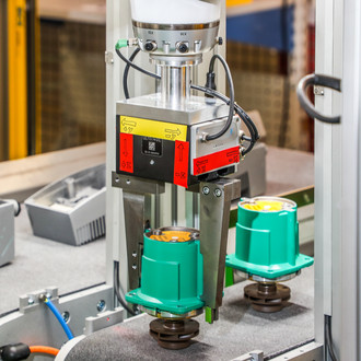 Foto einer industriellen Maschine, die zwei grüne Bauteile auf einem Förderband bearbeitet. Die Maschine hat eine metallene Haltevorrichtung mit Sensoren und verschiedenen Kabeln. Im Hintergrund sind weitere industrielle Geräte und eine strukturierte Umgebung zu erkennen. Die Bauteile auf dem Förderband sind symmetrisch angeordnet und werden präzise von der Maschine positioniert.