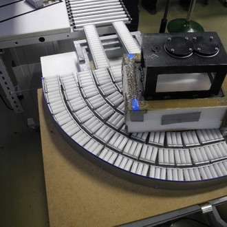 Foto einer industriellen Förderanlage, die aus mehreren gebogenen und geraden Rollenbahnen besteht. Die Förderanlage transportiert eine rechteckige schwarze Box, die mit Klebeband und anderen provisorischen Materialien befestigt ist. Die gebogenen Rollenbahnen formen einen Viertelkreis, der die Box um eine Kurve führt. Im Hintergrund sind weitere Teile der Anlage und der Fabrikboden zu sehen. Die Szene vermittelt einen Eindruck von einem Produktionsprozess mit modularen und anpassbaren Förderkomponenten.