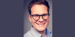 Foto eines lächelnden Mannes mit kurz geschnittenem Haar und Brille. Er trägt ein hellblaues Hemd und schaut direkt in die Kamera. Der Hintergrund ist dunkel und neutral gehalten, wodurch der Fokus auf dem Gesicht des Mannes liegt.