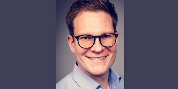 Foto eines lächelnden Mannes mit kurz geschnittenem Haar und Brille. Er trägt ein hellblaues Hemd und schaut direkt in die Kamera. Der Hintergrund ist dunkel und neutral gehalten, wodurch der Fokus auf dem Gesicht des Mannes liegt.