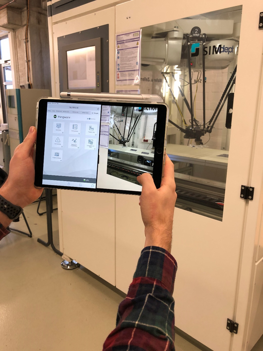 Foto: Eine Person hält ein Tablet, auf dem eine Bedienoberfläche für eine Maschine angezeigt wird. Die Person steht vor einer großen, weißen Maschine mit einem Fenster, durch das Teile eines robotischen Systems sichtbar sind. Auf dem Tablet ist auch ein Bild der Maschine zu sehen, die im Hintergrund steht. Die Person trägt ein kariertes Hemd und bedient das Tablet offensichtlich, um die Maschine zu steuern oder zu überwachen. Im Hintergrund sind weitere industrielle Geräte und ein heller Arbeitsbereich zu erkennen.