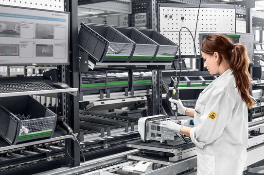 Das Bild zeigt eine Frau in einem weißen Laborkittel, die in einer modernen Produktionsumgebung arbeitet. Sie steht an einem Montageband und verwendet ein Handwerkzeug, um ein Bauteil zusammenzusetzen. Links von ihr sind mehrere Regale mit schwarzen Behältern zu sehen, die verschiedene Kleinteile und Materialien enthalten. Ein Monitor an der linken Seite des Bildes zeigt Anweisungen oder Diagramme, die den Montageprozess unterstützen. Die Umgebung ist sauber und organisiert, und die Frau trägt weiße Handschuhe, was auf eine präzise und saubere Arbeitsumgebung hinweist.