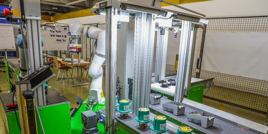 Foto einer automatisierten Produktionslinie in einer Fabrikhalle. Im Vordergrund befinden sich grüne Maschinen mit einem Förderband, auf dem mehrere kleine, runde Bauteile angeordnet sind. Links im Bild ist ein Roboterarm zu sehen, der offenbar in den Produktionsprozess eingebunden ist. Im Hintergrund sind Gitterabtrennungen und Tische mit Dokumenten sowie weitere industrielle Geräte erkennbar. Die Szene ist gut beleuchtet und deutet auf eine moderne Fertigungsumgebung hin.