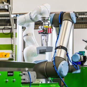 Foto von zwei Roboterarmen in einer industriellen Umgebung. Im Vordergrund befindet sich ein grauer Roboterarm mit blauen Kabeln, der zur Seite zeigt. Im Hintergrund ist ein weißer KUKA-Roboterarm, der ein Greifwerkzeug hält und gerade ein graues Bauteil manipuliert. Um die Roboter herum sind grüne Maschinen und Arbeitsflächen zu sehen. Die Szene zeigt die Anwendung fortschrittlicher Robotik in einer modernen Produktionslinie.