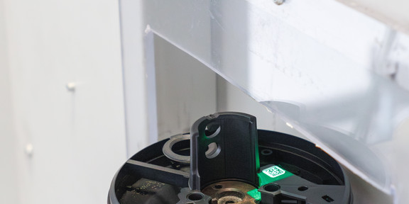 Nahaufnahme eines technischen Prüfgeräts in einem Labor: Ein rundes schwarzes Bauteil liegt eingespannt auf einer Metallplatte, daneben ist ein kleiner grüner Sensor mit Licht zu sehen