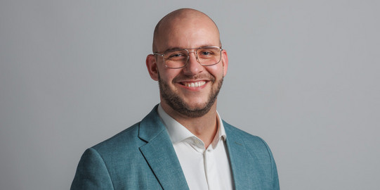 as Bild zeigt ein Porträtfoto eines lächelnden jungen Mannes mit kurzem, dunklem Haar und Brille. Er trägt ein schwarzes Sakko, ein weißes Hemd und eine graue Krawatte. Der Hintergrund ist weiß und zeigt die Buchstaben "IPS" in großen, grünen und grauen Blockbuchstaben. 