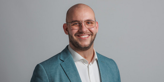 as Bild zeigt ein Porträtfoto eines lächelnden jungen Mannes mit kurzem, dunklem Haar und Brille. Er trägt ein schwarzes Sakko, ein weißes Hemd und eine graue Krawatte. Der Hintergrund ist weiß und zeigt die Buchstaben "IPS" in großen, grünen und grauen Blockbuchstaben. 
