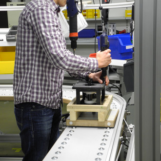 Ein Mann in einem karierten Hemd steht an einem Förderband und verwendet ein Elektrowerkzeug, um eine Baugruppe zusammenzusetzen. Im Hintergrund befinden sich gelbe und blaue Behälter mit verschiedenen Bauteilen.