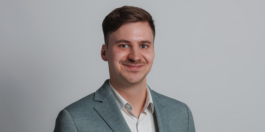 Das Bild zeigt einen jungen Mann mit kurzen braunen Haaren, der einen dunklen Anzug, ein weißes Hemd und eine gestreifte Krawatte trägt. Er lächelt in die Kamera. Im Hintergrund sind große grüne Buchstaben „IPS“ zu sehen, die teilweise durch den Mann verdeckt werden. Der Hintergrund ist schlicht und weiß gehalten.