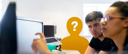 Zwei Personen sitzen vor einem Computerbildschirm und diskutieren etwas auf dem Bildschirm. Eine dritte Person ist als Silhouette dargestellt, die in orange und mit einem Fragezeichen versehen ist, was auf eine noch nicht besetzte Position hinweisen könnte. Im Hintergrund sind weitere Computer und ein Fenster zu sehen, das Tageslicht in den Raum lässt.